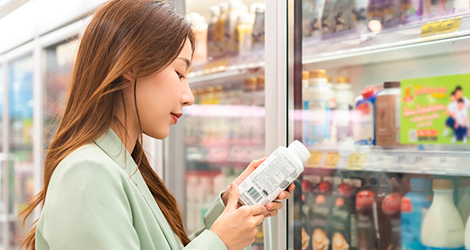 Mulher analisando o rótulo da embalagem de um produto no supermercado. Ela é branca, veste uma camisa verde e segura o produto nas mãos. 